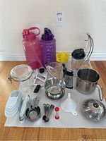 Photo of two large flip lid water bottles in pink and purple, yellow citrus juicer, Ninja Bullet mug, stainless steel pitcher, various measuring cups and spoons, grater, mandolin slicer, steamer basket, funnel, sealed jar, and egg timer spread on white surface.