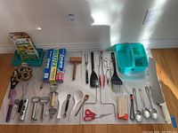 Wide view of all kitchen utensils and tools laid out including napkin rings, peelers, cooking spoons, tongs, scissors, mallet, waxed paper, and plastic caddy