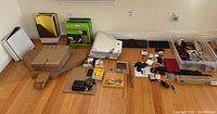 Wide view of office supplies, electronics, and accessories arranged on the floor, including binders, keyboards, clocks, cables, scissors, and boxes