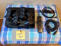 Top view of PS3 console with four black controllers laid on top, Sony remote, HDMI and AV cables to the right on a blue checkered cloth.