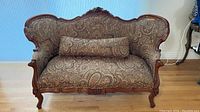 Front view of camel back settee with paisley upholstery, wood frame and bolster pillow.
