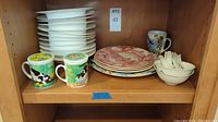Shelf with stacked white soup bowls, 4 red pattern plates, 2 covered tea mugs with strainers featuring cow designs, bluebird mug, small bowls, and ceramic spoons.