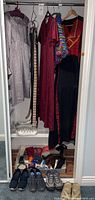 Photo of multiple women's dresses hanging including silver-gray, burgundy and black with red pattern dresses with leather belts and accessories placed on the floor below and shoes.