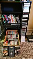 Photo of shelving unit with various VHS tapes, Sony disc player, and a box filled with numerous CDs stacked inside and additional CDs on floor in front.