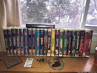 Full view of 23 Disney VHS tapes on a wooden windowsill, one Fantasia tape laid atop the row; three glass sun catchers and an empty DVD case in front; snowy yard visible outside.
