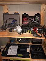 View of two corded drills, drill bit sets in plastic cases, Porta Fix 37-piece repair kit, and Mastercraft Maximum ratchet set on wooden shelf.