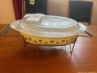 Side profile view of the Pyrex casserole dish with snowflake pattern in yellow and black, covered with the clear lid on the metal serving stand.