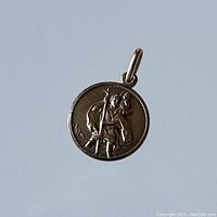 Front view of small circular gold pendant showing raised relief of a walking figure with a staff and cloak.