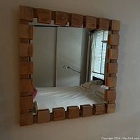 Full view of the square mirror hung on the wall showing the wooden block frame pattern and reflection of a room interior.