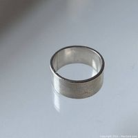 Sterling silver band ring shown on reflective surface, highlighting the brushed finish and width.