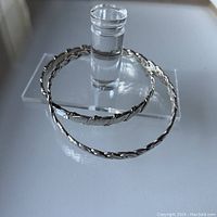 Two sterling silver bangles with twisted and wave designs displayed on clear stand.