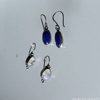 Photo of two pairs of earrings on a reflective white surface, one pair with blue sodalite stones and one pair with white moonstones.