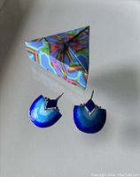 Pair of sterling silver enamel earrings with blue gradient drop and diamond shaped top accents next to colorful origami-style box