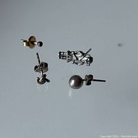 Four stud earrings arranged on a reflective surface: small round sterling silver stud with black onyx inset, silver knot stud, silver ball stud, and pewter figurine stud.