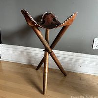 Front view of folding stool with three wooden legs and brown leather saddle seat, showing overall structure and wood grain.