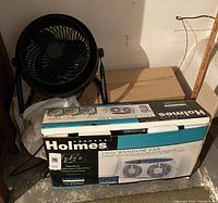 Photo showing a black standing fan positioned beside a boxed Holmes twin window fan in a corner with concrete floor and wall.
