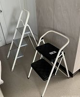 Two step stools: one three-step metal with white frame and handrail, and one two-step black steps with white frame and rounded top handrail, positioned next to a wall and door.