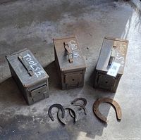 Three rectangular metal ammo boxes with handles on top, placed on concrete floor with four rusty horse shoes in front. Ammo boxes are marked with white paint and show signs of rust and wear. The horse shoes vary in size and are rusty.