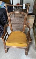Antique carved rattan chair showing woven backrest, carved top panel, armrests, and mustard seat cushion with stain.