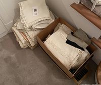 Photo of stacked vintage linens including pillow cases and a box of lace tablecloths and napkins on carpeted floor beside stairs.
