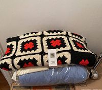 Two folded textiles stacked, the top is a hand knit afghan with black, white, and red squares. The bottom is a light blue blanket in protective packaging.