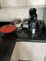 Photo shows six small kitchen appliances on black countertop: electric wok, rice cooker, blender, toaster, coffee maker, and a small plastic container.