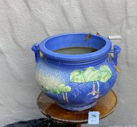 Image showing a large blue pottery planter with raised green tree designs sitting on a small round wooden table against a textured wall. The planter is round with a bulbous body, two looped handles, and a wide opening.