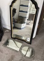 Large wall mirror standing vertically with black and gold scalloped frame. Two smaller mirrored mats placed on the floor in front of it: a rectangular one with floral etching and a circular one with floral etching separated into two halves.