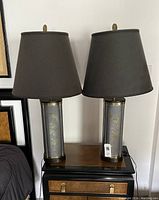 Pair of dark grey metal Asian style table lamps with brass overlay designs on bases and dark brown fabric lampshades sitting on a wooden surface against a wall.