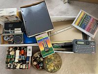 Wide view of entire lot showing label makers, colored pencils, thread spools, buttons, sheet protectors and other office supplies in a box