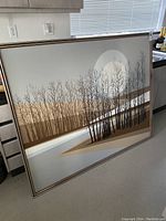 Large framed abstract landscape painting featuring bare trees in earth tones and metallic background, placed leaning against kitchen cabinets.