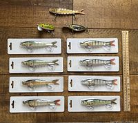 All 11 fishing lures arranged flat on wood surface with ruler for scale, including 8 new lures in original white plastic packaging and 3 unpackaged lures.