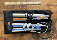 Two hair styling tools: a hair crimper with silver and orange plates and a hair straightener in white and blue color, placed on a black drawstring bag.