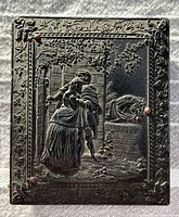 Close-up photo of the box lid with detailed embossed scene of man and woman, floral border, and visible cracks/chips on dark stone-like surface