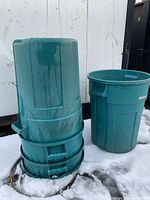 Four teal-colored plastic outdoor trash cans stacked and displayed on snow against a white wall. Two cans are stacked upside down, and one is upright showing the handles.