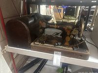 Antique Singer sewing machine on a metal shelf, showing the entire machine mounted on a wooden base with attached electric components and a curved wooden case nearby