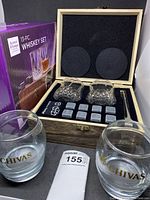 Photo showing open wooden box with two crystal cut glasses, whiskey stones, tongs, funnel, alongside the closed packaging box and two Chivas branded glasses in front.