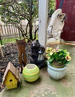 View of metal cat figurine, bear figurine holding lantern, small round ceramic planter, yellow wooden birdhouse, larger floral ceramic planter, and white Toro leaf bag on ground