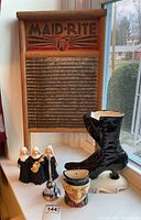 Photo of Maid-Rite washboard, black ceramic Victorian-style boot, three musical nun figurines, silver-tone bell, character face mug, and white pig figurine arranged on a windowsill.