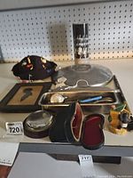 Several vintage collectibles arranged on a ledge against pegboard. Includes felt pincushion, metal tray, framed silhouette, ceramic duck, glass-topped box, metal cup.