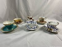 Photo of all six English china teacups with saucers arranged and displayed on white fabric background showing variety of patterns and colors.