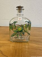 Front view of clear glass decanter with painted green fruit and leaf design on wooden surface.