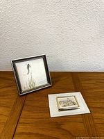 Wide view showing both small framed artworks on wooden table.