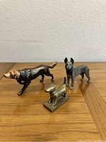 Front and side angle of three metal dog figurines on wooden surface against wall.