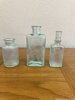 Three vintage glass bottles of varying shapes and sizes displayed on a wooden table against a beige wall, showing clear detail of bottle features and slight aqua tint.