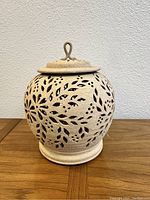 Front view of the round stone lattice pot with visible cut-out floral patterns and detachable lid with loop handle.