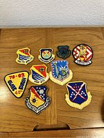 Nine vintage military patches laid out on a wooden surface showing various colors and unit insignias.