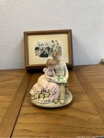 Porcelain figurine of boy and girl sitting on a base, with girl in pink dress holding flowers and boy reading a book, placed in front of framed vintage artwork.