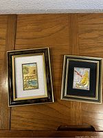 Two framed artworks on a wooden table, showing both the landscape and sailboat images, captured from a slight angle.