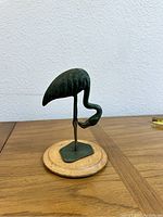 Side profile view of green flamingo statue on wooden round base placed on wooden surface against a white wall.